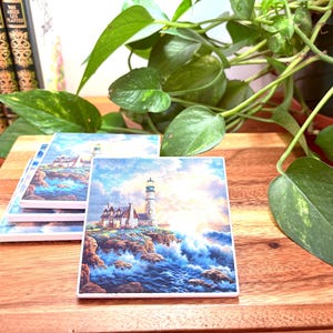 May include: Square ceramic coasters featuring a lighthouse scene. The image shows a white lighthouse on a cliff above a rough sea. The sky is a blend of blue and white, with bright light breaking through the clouds. The coasters are stacked on a wooden surface.
