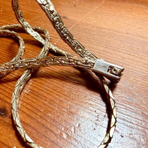 May include: Close-up of a gold-toned necklace with a woven chain design. The clasp is silver-colored and marked with "18K". The necklace is displayed on a wooden surface.