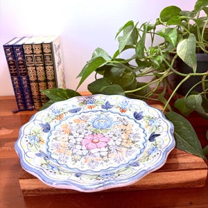 May include: A decorative ceramic plate with a scalloped edge. The plate features a floral design with pink, blue, yellow, and orange flowers. The rim is light blue. The plate is on a wooden surface.