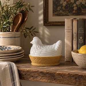 May include: A white milk glass hen on a nest dish featuring a detailed chicken figurine lid resting on a yellow, basket-weave textured base. The piece is a decorative container designed with moulded feather patterns on the hen and a woven design.