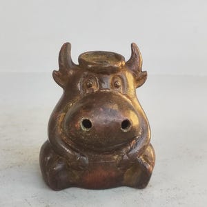 May include: A bronze-colored, seated cow figurine with a whimsical design. The cow has large, round eyes, a prominent snout with two nostrils, and small horns. The figurine is made of metal and has a weathered appearance.