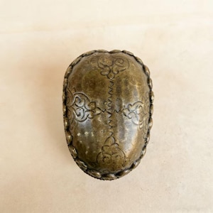 May include: An antique brass-colored container with an oval shape. The surface is adorned with intricate floral and scrollwork patterns. The edges are detailed with a beaded design, adding to its vintage aesthetic.