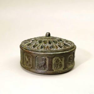 May include: A bronze-colored, round trinket box with a decorative, pierced lid and a small knob. The sides feature embossed panels with animal figures. The box has a weathered appearance, suggesting age or a vintage style.