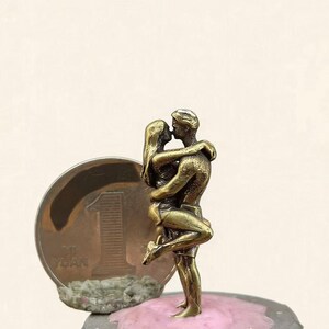Puede incluir: Peque&ntilde;a escultura de bronce de una pareja abraz&aacute;ndose y bes&aacute;ndose. Las figuras son detalladas y se encuentran sobre una superficie rosa encima de una chapa de botella. Se ve una moneda con el n&uacute;mero 1.