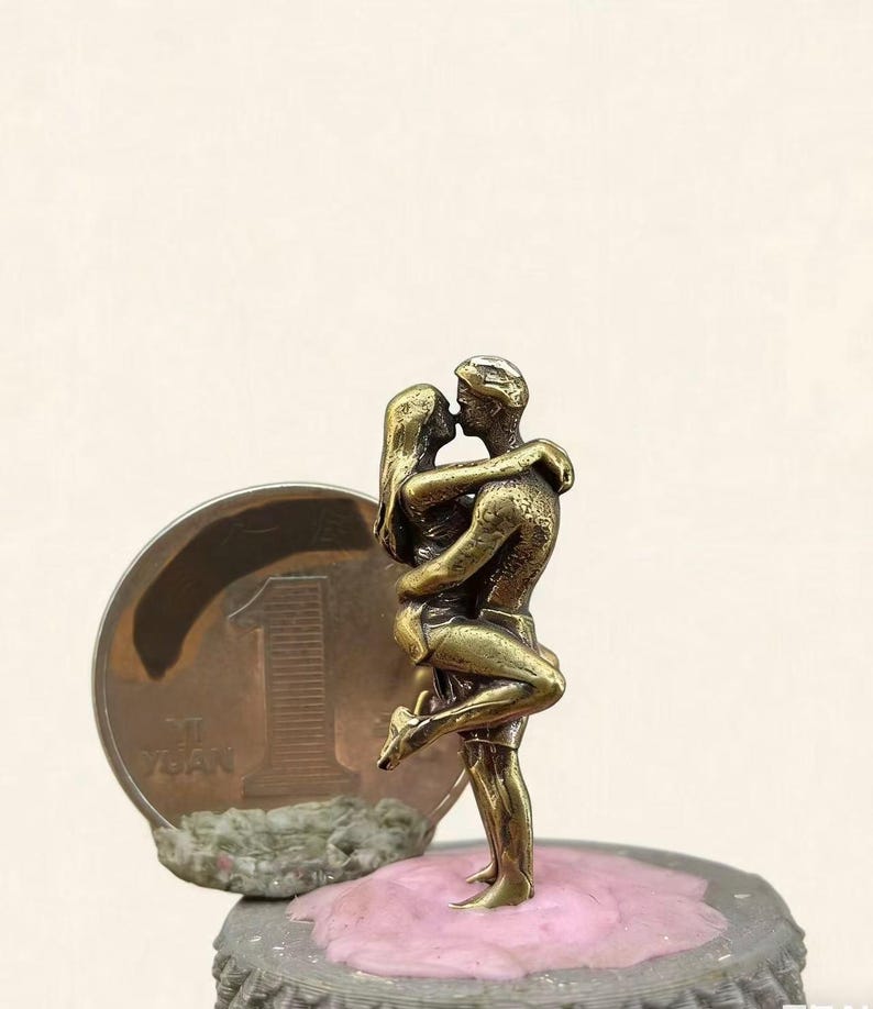 Puede incluir: Peque&ntilde;a escultura de bronce de una pareja abraz&aacute;ndose y bes&aacute;ndose. Las figuras son detalladas y se encuentran sobre una superficie rosa encima de una chapa de botella. Se ve una moneda con el n&uacute;mero 1.