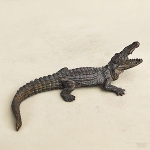 May include: A bronze-effect alligator figurine with an open mouth, showing detailed teeth. The reptile has a textured, scaled body and is posed on a neutral background. The figurine is a decorative item.