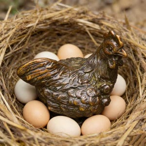 Può includere: Una statuetta di gallina color bronzo, adagiata in un nido di paglia, circondata da una dozzina di uova color crema e marrone. La gallina presenta dettagli di piume e una cresta prominente. Un oggetto decorativo rustico.