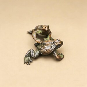 以下が含まれることがあります： 2匹のカエルのブロンズ彫刻。1匹がもう1匹の上にいます。カエルは質感のある肌と緑がかった茶色のパティナを持っています。背景はニュートラルカラーです。