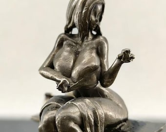 May include: A metallic silver-toned figurine depicting a seated figure with long hair, draped in a cloth covering the lower body. The figure is positioned with one arm extended outward and the other hand resting near the chest area, finished in a textured, monochromatic metallic surface.