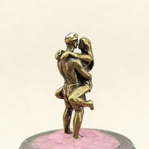Puede incluir: Una escultura de bronce de una pareja abraz&aacute;ndose y bes&aacute;ndose. El hombre sostiene a la mujer, y est&aacute;n sobre una base rosa y gris. La escultura es detallada, representando la ropa y los rasgos de las figuras.