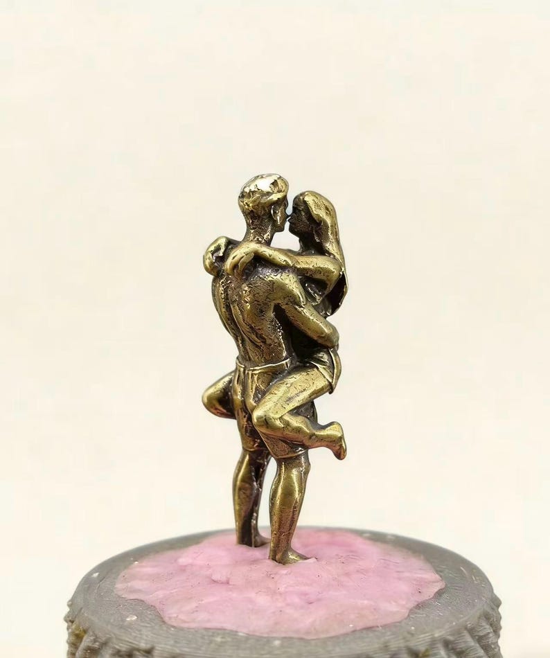 Puede incluir: Una escultura de bronce de una pareja abraz&aacute;ndose y bes&aacute;ndose. El hombre sostiene a la mujer, y est&aacute;n sobre una base rosa y gris. La escultura es detallada, representando la ropa y los rasgos de las figuras.