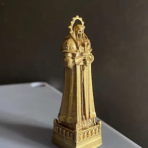 May include: A gold-colored statue of a robed figure, possibly a saint or religious figure, holding objects. The statue has a halo and is standing on a detailed base. The figure is wearing a long robe.