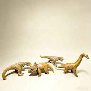 Pode incluir: Quatro dinossauros de brinquedo em tons de bege e castanho estão dispostos sobre um fundo claro. A coleção inclui um Tyrannosaurus Rex, um Triceratops e um Brachiosaurus, entre outros. Estas figuras de plástico são detalhadas e texturizadas.