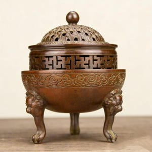 May include: A bronze incense burner with a domed lid featuring intricate cut-out patterns. The burner has a decorative band with a geometric design and rests on three lion-paw feet. The overall design is ornate and detailed.