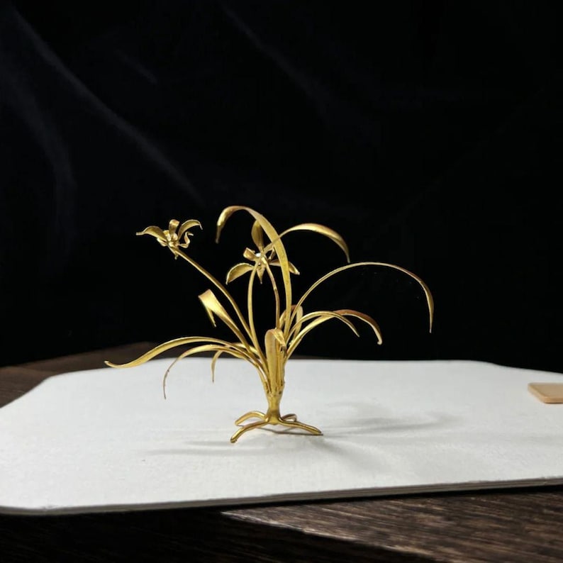 May include: A golden metal sculpture of a plant with long, slender leaves and delicate flowers. The sculpture is set against a white surface with a dark background, highlighting the intricate details of the handcrafted piece.