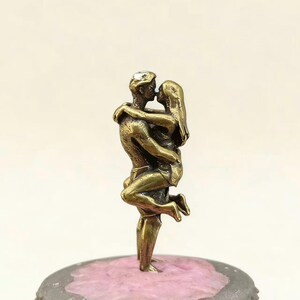 Puede incluir: Una escultura de color bronce de una pareja abraz&aacute;ndose y bes&aacute;ndose. El hombre levanta a la mujer y sus cuerpos est&aacute;n entrelazados. La escultura est&aacute; montada sobre una base gris con una superficie rosa.
