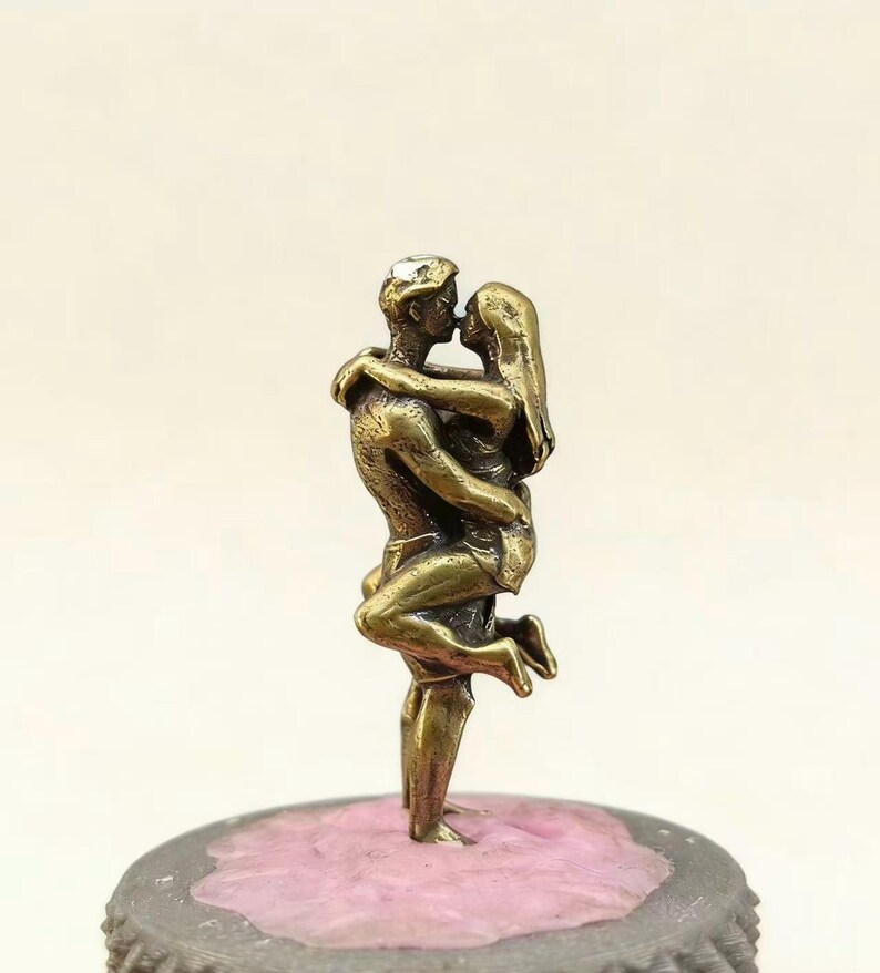 Puede incluir: Una escultura de color bronce de una pareja abraz&aacute;ndose y bes&aacute;ndose. El hombre levanta a la mujer y sus cuerpos est&aacute;n entrelazados. La escultura est&aacute; montada sobre una base gris con una superficie rosa.