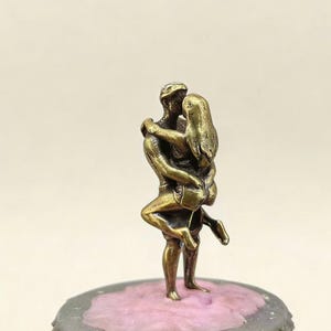 Puede incluir: Peque&ntilde;a escultura de bronce que representa a una pareja abraz&aacute;ndose. El hombre sostiene a la mujer en sus brazos, sus cuerpos entrelazados. La escultura est&aacute; montada sobre una base rosa y gris.
