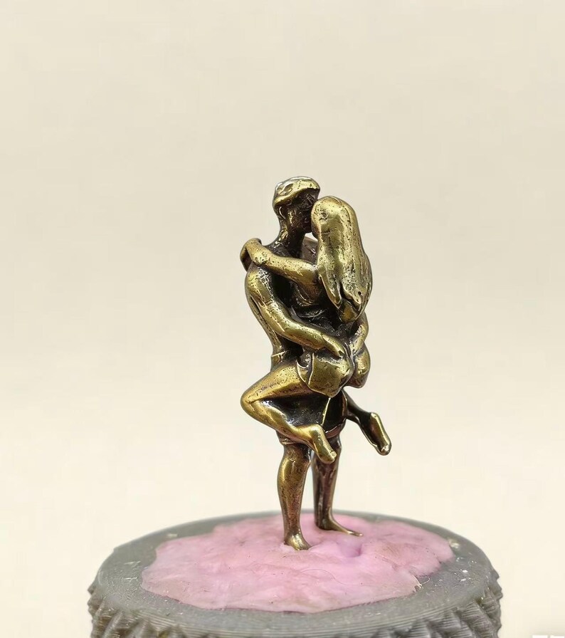 Puede incluir: Peque&ntilde;a escultura de bronce que representa a una pareja abraz&aacute;ndose. El hombre sostiene a la mujer en sus brazos, sus cuerpos entrelazados. La escultura est&aacute; montada sobre una base rosa y gris.
