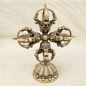 Puede incluir: Un Vajra detallado de color bronce, un objeto ritual, con intrincados diseños florales y geométricos. El objeto tiene forma de cruz con brazos curvos y una base decorativa. El diseño general es simétrico y ornamentado.