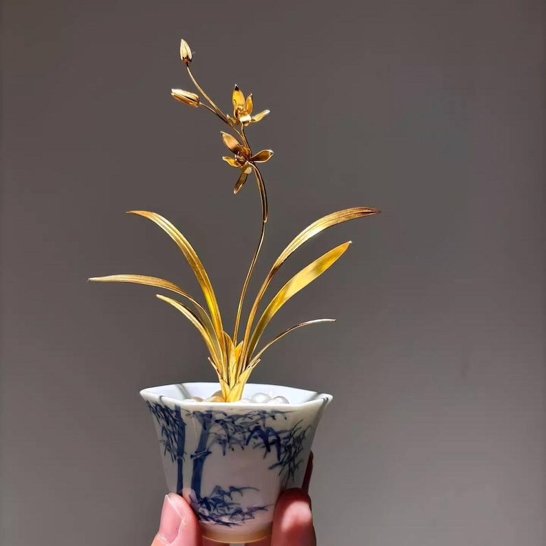 May include: A golden metal orchid sculpture in a small, white and blue porcelain pot. The pot features a bamboo design. The orchid has detailed petals and leaves, and is held in a hand against a gray background.