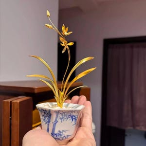 May include: A golden orchid sculpture in a white and blue porcelain pot. The sculpture features detailed leaves, stems, and flowers. The pot has a blue bamboo design. The sculpture is held in a hand.