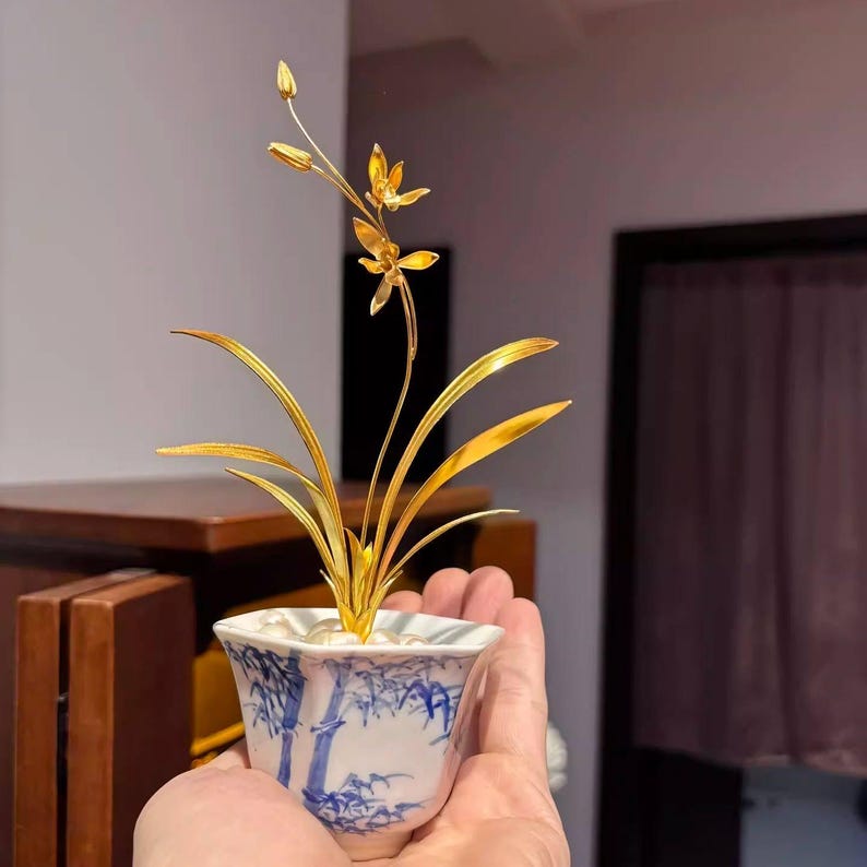 May include: A golden orchid sculpture in a white and blue porcelain pot. The sculpture features detailed leaves, stems, and flowers. The pot has a blue bamboo design. The sculpture is held in a hand.