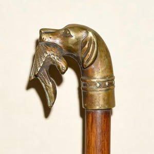 May include: Antique walking cane with a brass dog head handle holding a bird in its mouth. The handle has a detailed design and is attached to a wooden shaft. The cane is a vintage item.