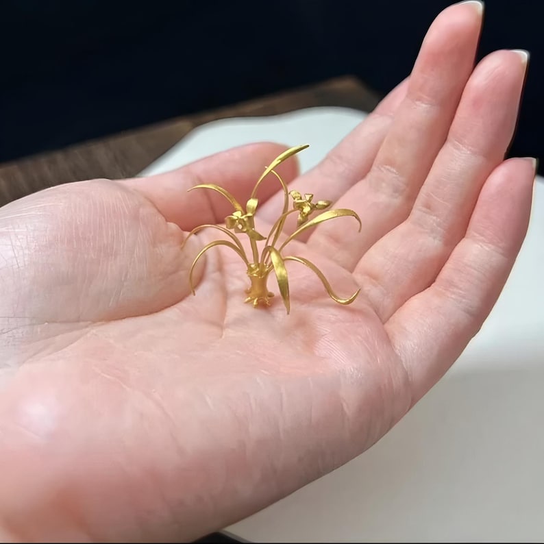 May include: A small, gold-colored floral sculpture featuring delicate leaves and flowers. The sculpture is held in a hand, showcasing its intricate details and craftsmanship. The piece has a matte finish and appears to be made of metal.