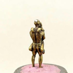 Puede incluir: Peque&ntilde;a escultura de color bronce que representa dos figuras abraz&aacute;ndose. Las figuras est&aacute;n estilizadas con detalles, de pie sobre una base rosa y gris. La escultura tiene una est&eacute;tica vintage.