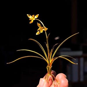 May include: A golden metal sculpture of a flower with two blooms and long, slender leaves. The sculpture is held against a dark background, highlighting its intricate details and metallic sheen. The flower's petals and leaves are delicately crafted.