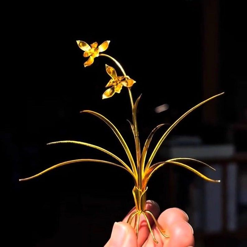 May include: A golden metal sculpture of a flower with two blooms and long, slender leaves. The sculpture is held against a dark background, highlighting its intricate details and metallic sheen. The flower's petals and leaves are delicately crafted.