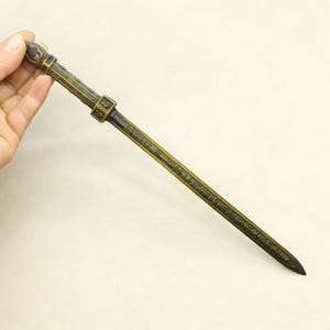 May include: A bronze-colored, ornate sword with a detailed handle and blade. The blade features engraved characters. The sword is held by a hand against a neutral background. The sword's overall design suggests a historical or cultural artifact.