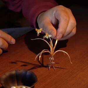 May include: A delicate, golden metal sculpture of a small plant with two flowers. The sculpture features thin, curved leaves and detailed roots. The plant is held by a person's hand against a dark wooden surface.