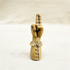 May include: A small, tan-colored sculpture of a hand making a gesture. The index and middle fingers are raised, while the other fingers are curled into the palm. The wrist area has a band around it, and there is text on the lower part of the sculpture.