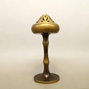 May include: A brass incense burner with a mushroom-shaped top featuring decorative cutouts. The burner has a slender, bamboo-like stem and a circular base. The overall design is a warm, antique gold color, suitable for home decor.