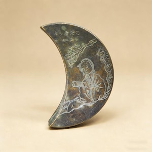 May include: A crescent-shaped, antique bronze object with etched artwork. The artwork depicts a figure in traditional clothing, possibly reading or writing, with surrounding foliage and text. The object has a weathered appearance, suggesting age and use.
