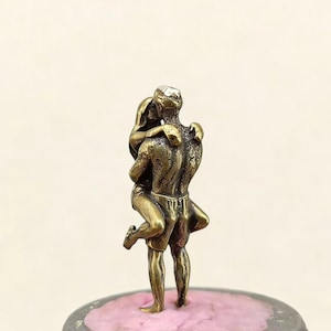 Puede incluir: Peque&ntilde;a escultura de color bronce que representa a una pareja abraz&aacute;ndose. El hombre levanta a la mujer, sus cuerpos entrelazados. La escultura est&aacute; sobre una base rosa texturizada, sugiriendo un tema rom&aacute;ntico.
