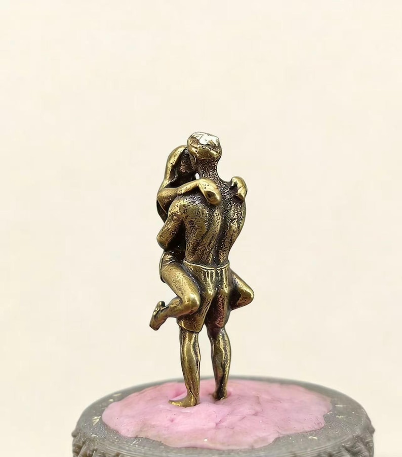 Puede incluir: Peque&ntilde;a escultura de color bronce que representa a una pareja abraz&aacute;ndose. El hombre levanta a la mujer, sus cuerpos entrelazados. La escultura est&aacute; sobre una base rosa texturizada, sugiriendo un tema rom&aacute;ntico.