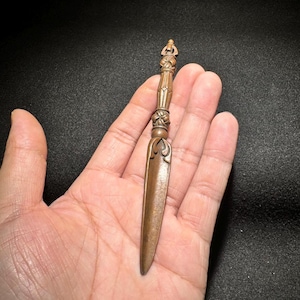 Könnte beinhalten: Ein bronzener Brieföffner mit aufwendigen Schnitzereien. Der Griff weist ein detailliertes Design mit einer kleinen Figur an der Spitze auf. Die Klinge ist lang und schlank, mit einem dekorativen Blumenmuster in der Nähe des Griffs.