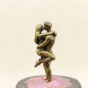 Puede incluir: Una escultura de color bronce de una pareja abraz&aacute;ndose y bes&aacute;ndose. El hombre levanta a la mujer, que se inclina hacia atr&aacute;s. La escultura est&aacute; sobre una base decorativa con una parte superior rosa.
