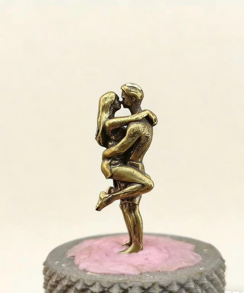 Puede incluir: Una escultura de color bronce de una pareja abraz&aacute;ndose y bes&aacute;ndose. El hombre levanta a la mujer, que se inclina hacia atr&aacute;s. La escultura est&aacute; sobre una base decorativa con una parte superior rosa.