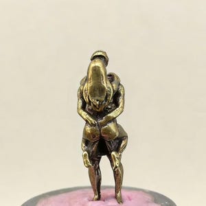 Puede incluir: Peque&ntilde;a escultura de color bronce que representa dos figuras abraz&aacute;ndose. Las figuras est&aacute;n entrelazadas, una parece estar cargando a la otra. La escultura est&aacute; montada sobre una base rosa y gris, mostrando un dise&ntilde;o detallado e &iacute;ntimo.