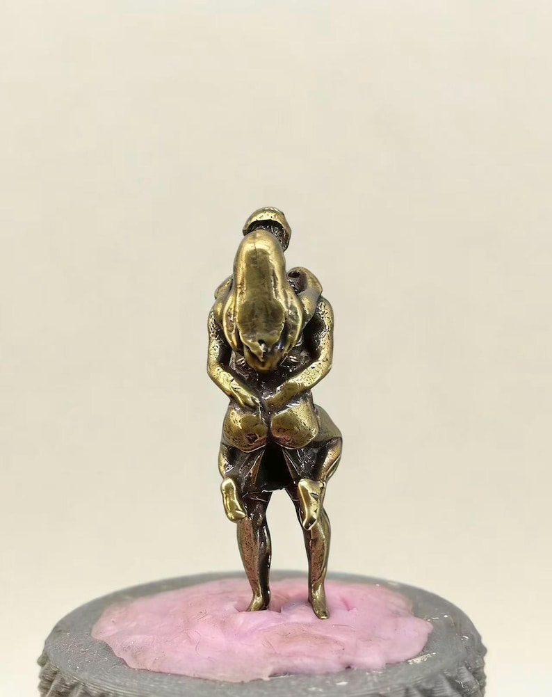 Puede incluir: Peque&ntilde;a escultura de color bronce que representa dos figuras abraz&aacute;ndose. Las figuras est&aacute;n entrelazadas, una parece estar cargando a la otra. La escultura est&aacute; montada sobre una base rosa y gris, mostrando un dise&ntilde;o detallado e &iacute;ntimo.