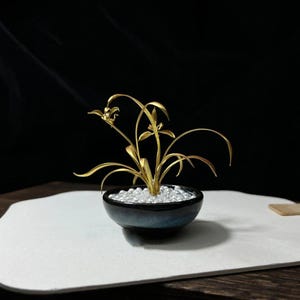 May include: A golden metal orchid sculpture in a small, dark blue ceramic pot filled with small, clear beads. The orchid has delicate leaves and flowers. The pot sits on a white surface against a black background.