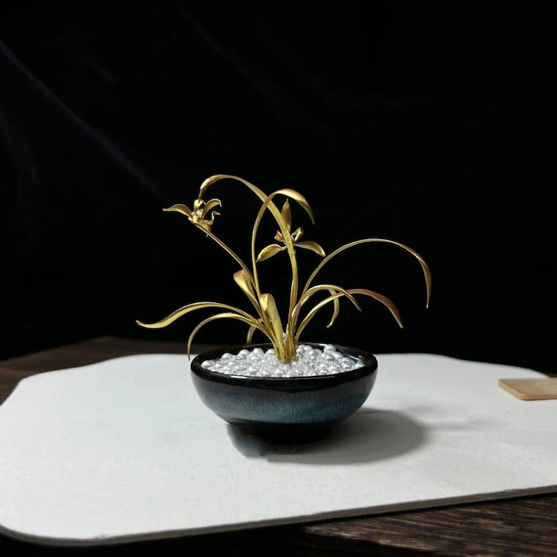May include: A golden metal orchid sculpture in a small, dark blue ceramic pot filled with small, clear beads. The orchid has delicate leaves and flowers. The pot sits on a white surface against a black background.
