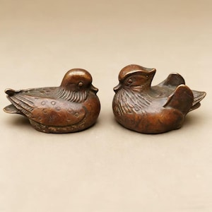 Könnte beinhalten: Zwei Entenfiguren aus Bronze. Eine Ente befindet sich in Ruheposition, die andere hat ihre Flügel ausgebreitet. Die Enten haben detaillierte Federmuster und sind warmbraun. Es sind wahrscheinlich Dekorationsartikel.