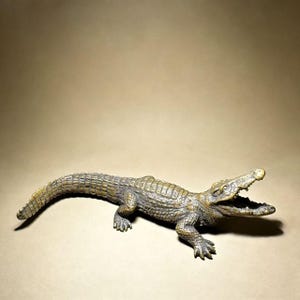 May include: A detailed, bronze-colored crocodile figurine with an open mouth, showcasing textured scales and realistic features. The sculpture is posed on a neutral background, highlighting its intricate design.