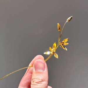 May include: A delicate, gold-colored floral hair accessory featuring a slender stem with two open flowers and two buds. The accessory is held against a gray background, showcasing its intricate design.