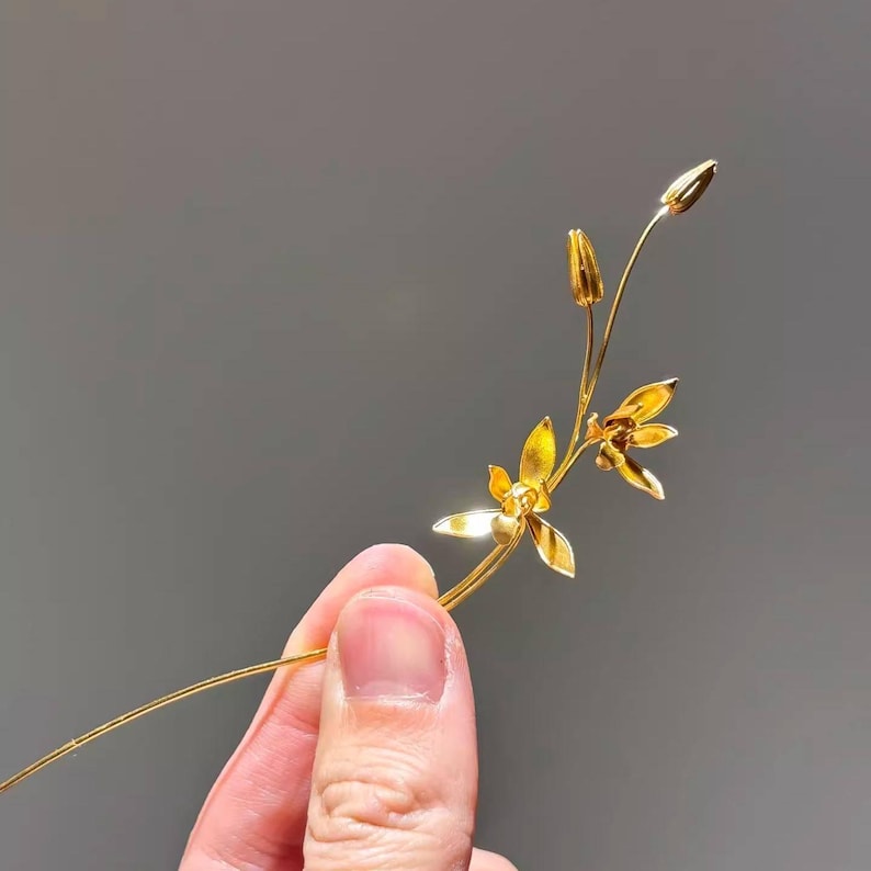 May include: A delicate, gold-colored floral hair accessory featuring a slender stem with two open flowers and two buds. The accessory is held against a gray background, showcasing its intricate design.